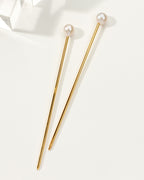 Two Ebb and Flow pearl hair pins, with gold shafts, lay on a white surface.