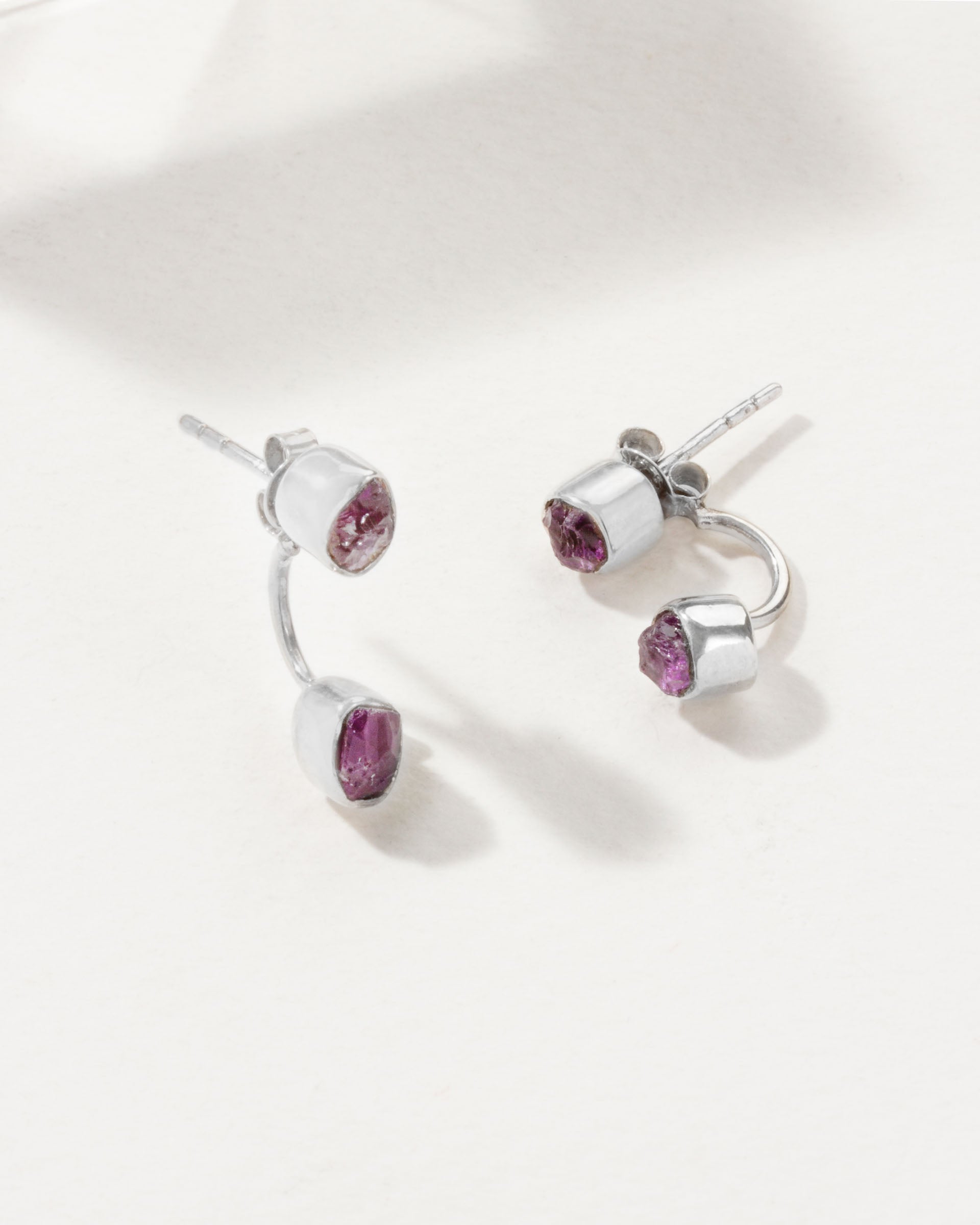 Two silver Double Trouble earrings with bezel set Amethyst gemstones, laying on a white surface.