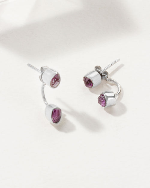 Two silver Double Trouble earrings with bezel set Amethyst gemstones, laying on a white surface.