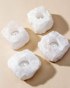 Four quartz candle holders with tea lights sit on a light tan surface.