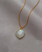 Moonstone heart-shaped pendant on a gold chain against a gray cloth background.