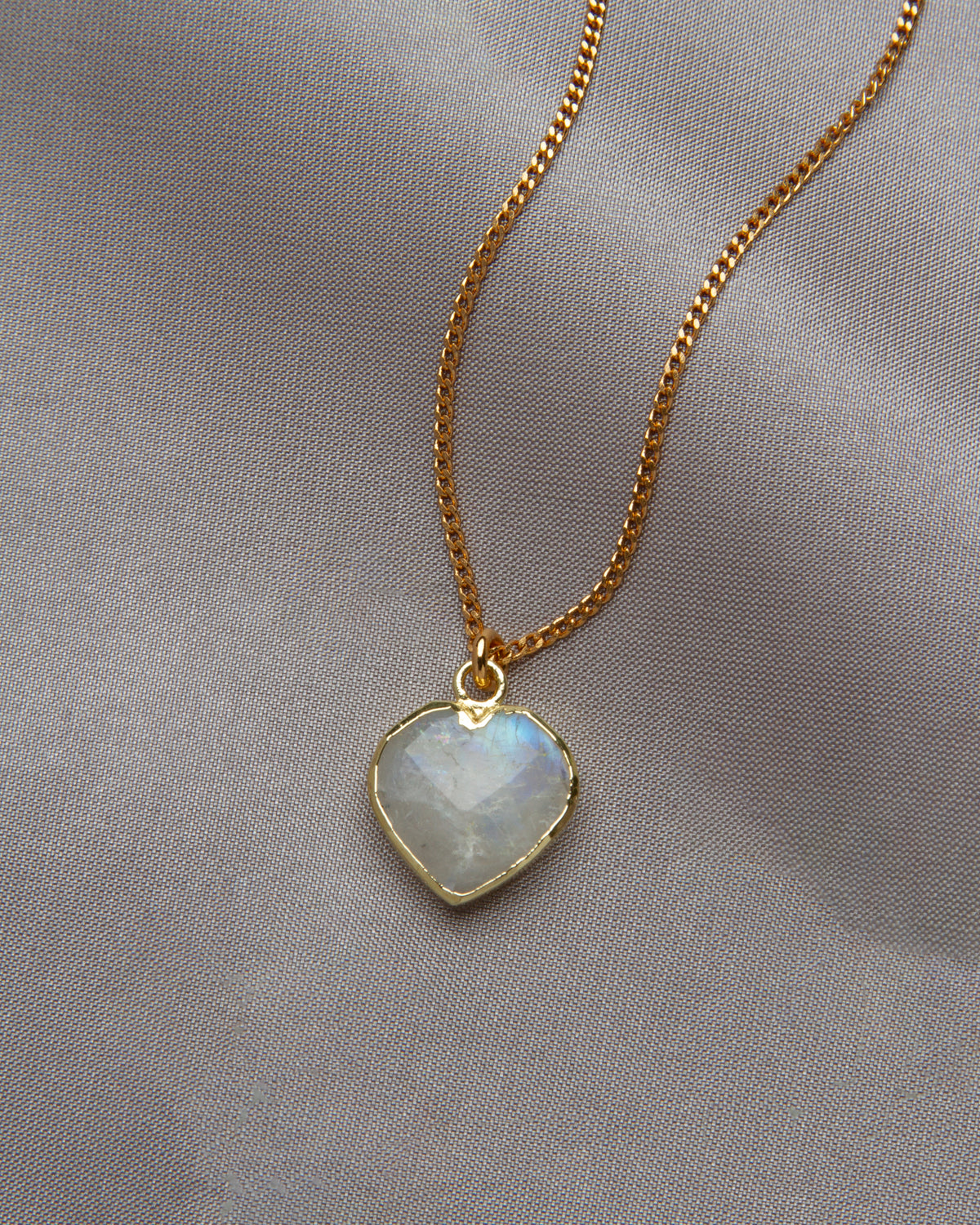 Moonstone heart-shaped pendant on a gold chain against a gray cloth background.
