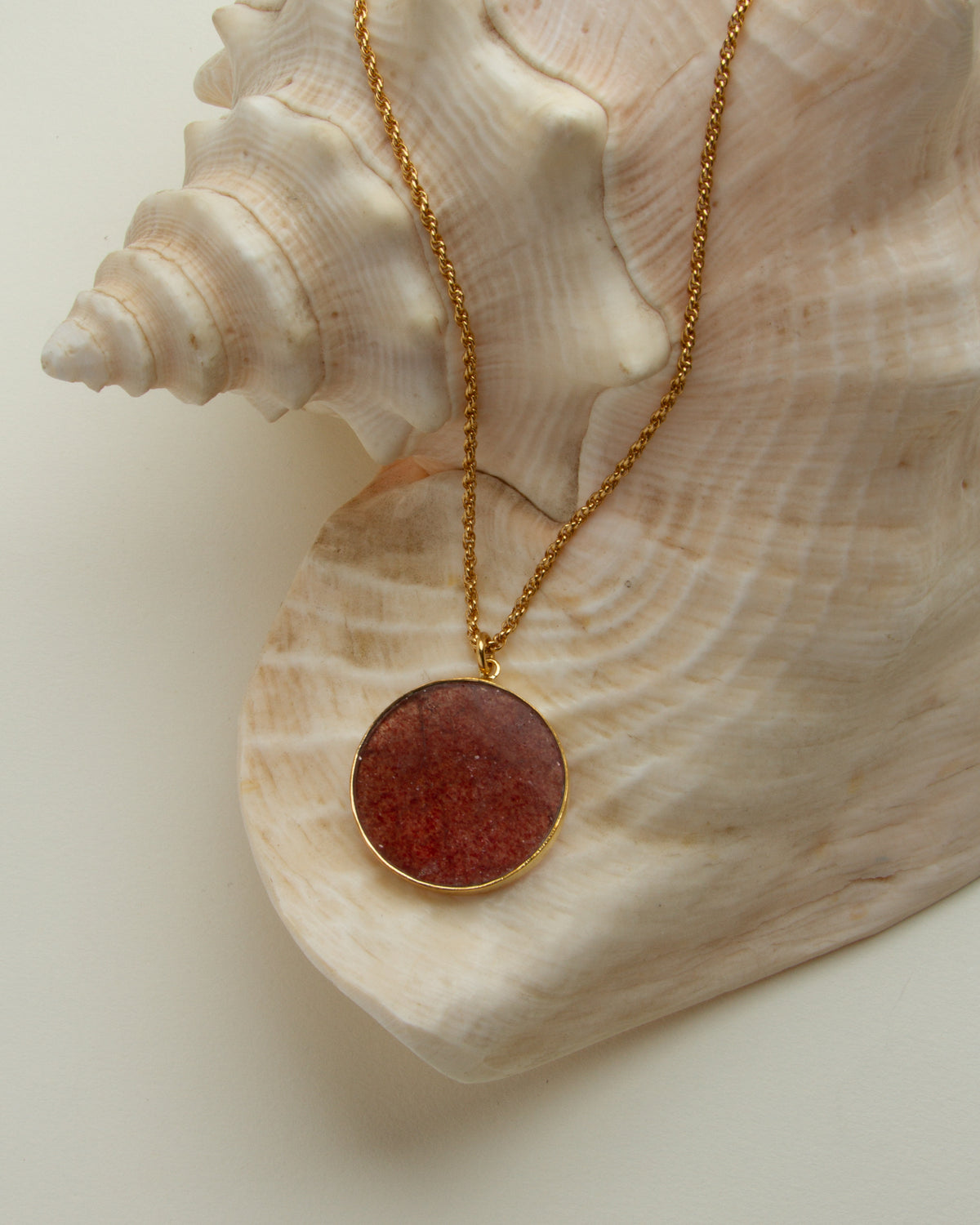 Gold chain necklace with strawberry quartz medallion against a seashell.