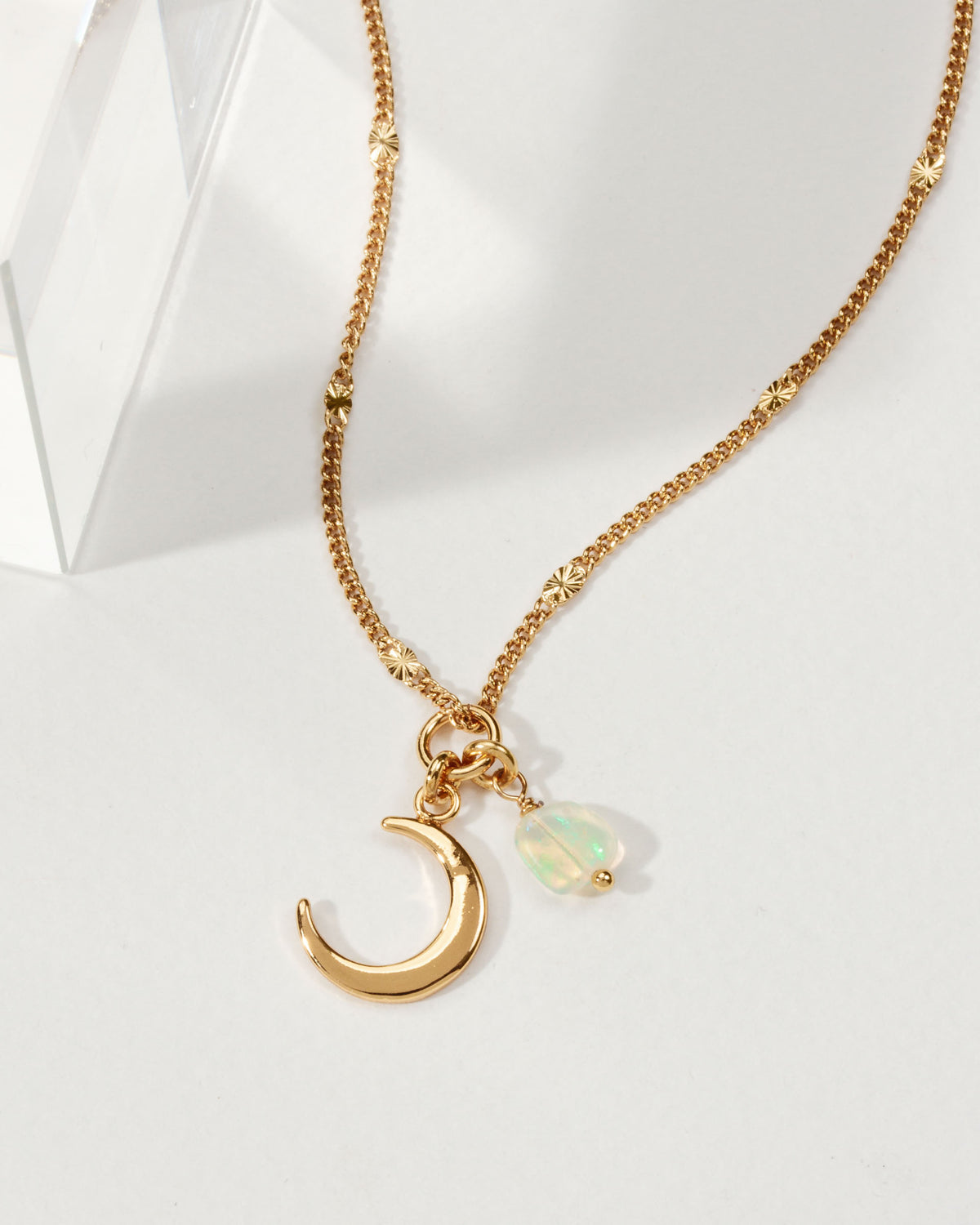 A gold chain necklace with a crescent moon and iridescent opal charm is displayed.