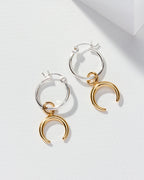 Two silver hoop earrings, each with a gold crescent charm, are displayed on a white surface.