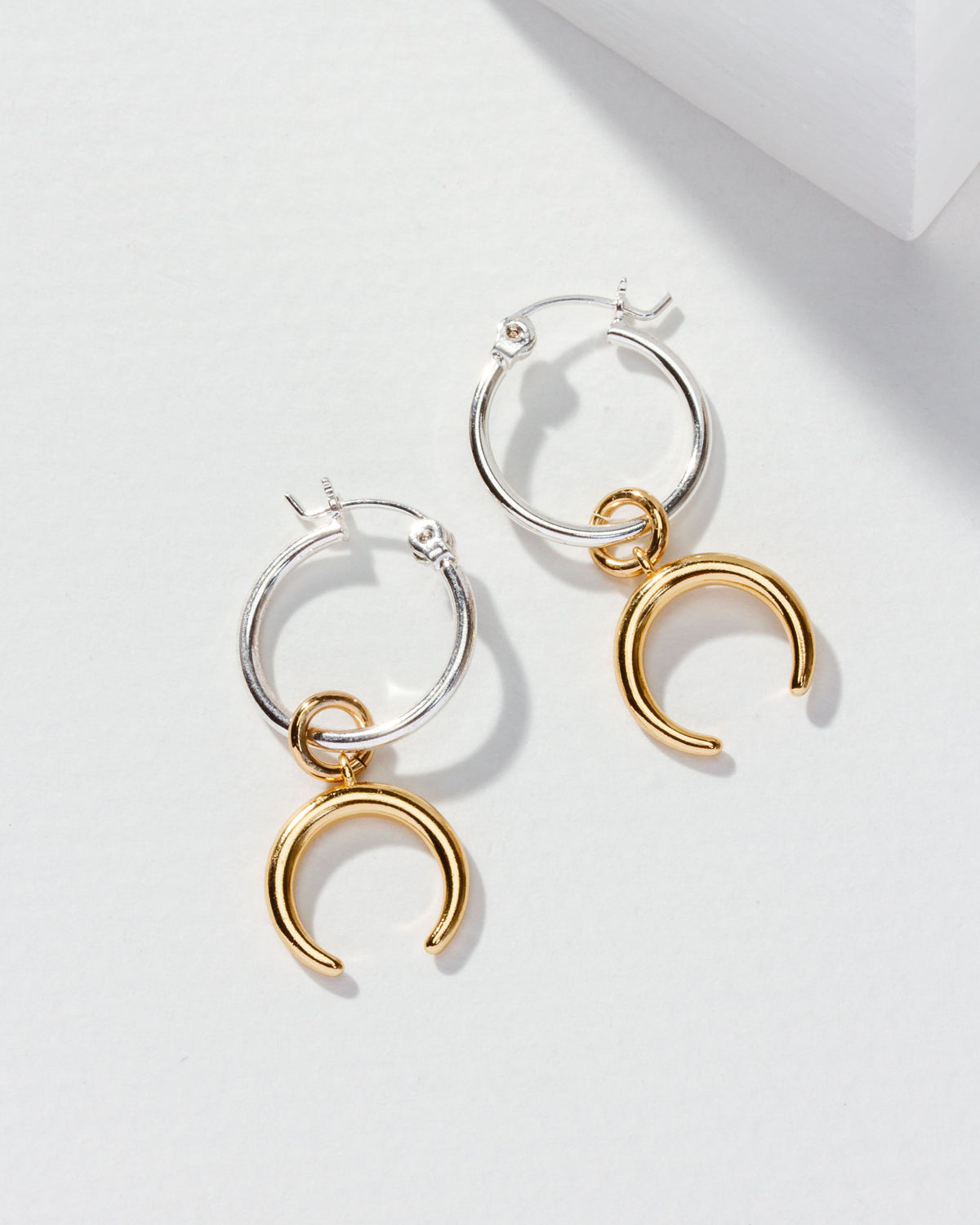 Two silver hoop earrings, each with a gold crescent charm, are displayed on a white surface.