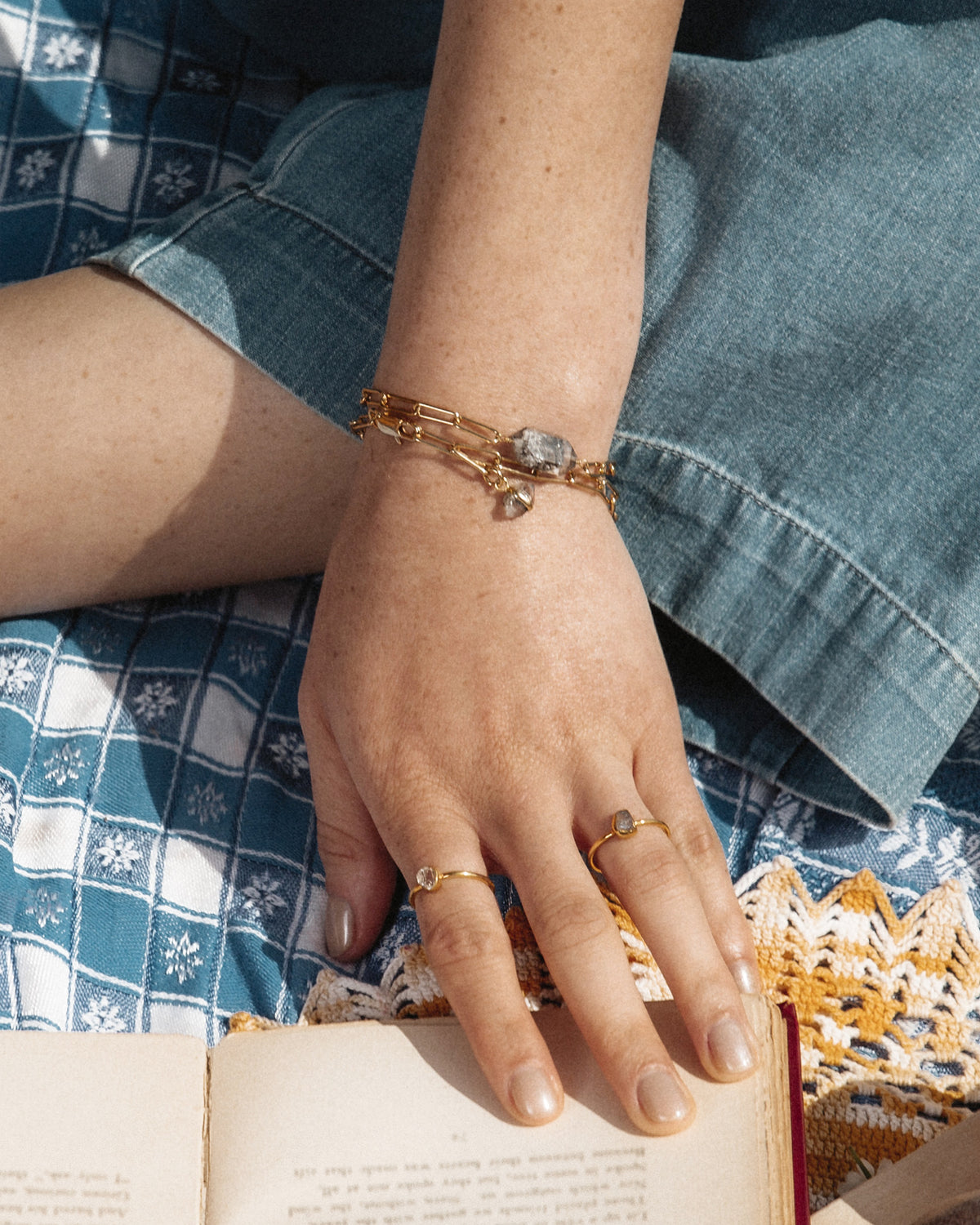 Hand wearing a Rock-On Bracelet with Herkimer Quartz stone and gold rings, resting on a book.