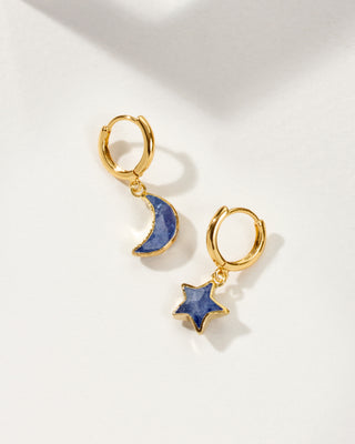 A pair of mismatched gold huggie hoop earrings with sapphire star and moon charms, set against a background of text.