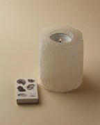 Selenite candle holder with lit candle next to a box of matches with shell images
