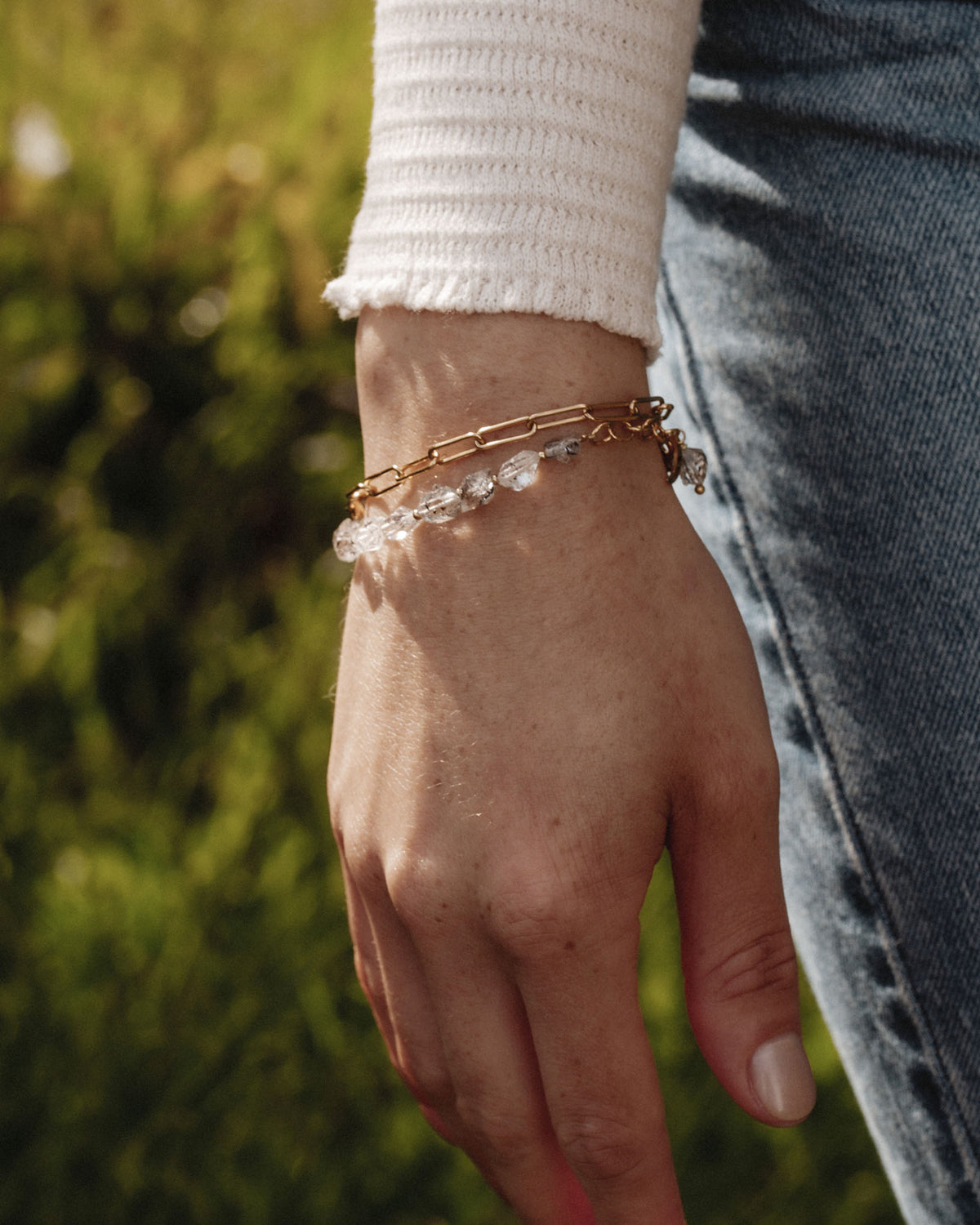 Hand wearing two bracelets; one gold chain and one with Herkimer Quartz gemstones.